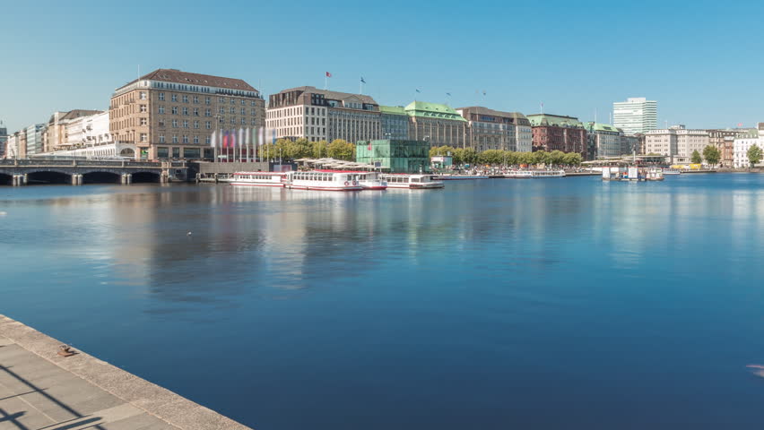 Panorama showing Binnenalster timelapse in Hamburg, Germany, with boat station. Historic buildings reflected in Inner Alster lake waters under a blue sky. Scenic urban landscape with swans at pier