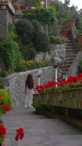 A woman in a white dress walks on a stone path in an Amalfi Coast garden, surrounded by greenery, flowers, lamp posts, and stone walls with potted plants.