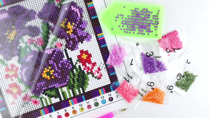 Assembled purple flower diamond mosaic fragment, tray with rhinestones, pen tool, rhinestones in transparent packaging and folded protective film, white background, close-up, top view, rotation, DIY