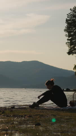 Lady throwing pebble stone into lake. Woman with food basket spends time enjoying moment of tranquility on riverbank at sunset. Weekend alone