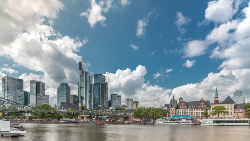 Hyperlapse of the historical Eiserner Steg (Iron Bridge) over the Main River in Frankfurt, Germany. Skyscrapers skyline and historic buildings reflect in the water under a cloud-filled sky timelapse