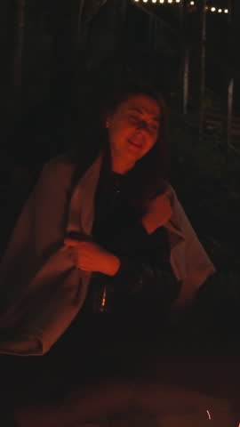 Young woman watches fire late at night. Wife adjusts blanket on shoulders sitting on stones by campfire. Time for diving in realm of dreams and aspirations