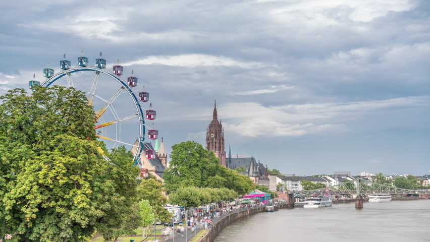 Aerial hyperlapse of pleasure boats on the Rhine Main, framed by the spire of Kaiserdom St. Bartholomaus. Eiserner Steg bridge and Ferris wheel add vibrancy to Frankfurt