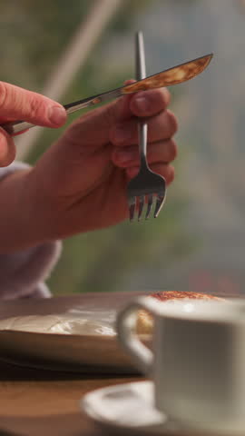 Man cuts piece of pancake at table in glamping house. Young man eats sweet breakfast against glass wall in eco-hotel. Standard room service breakfast