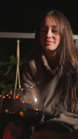 Wife cheers wineglass with husband on terrace. Young lady shares romantic candlelit dinner with partner in open-air restaurant among garlanded trees