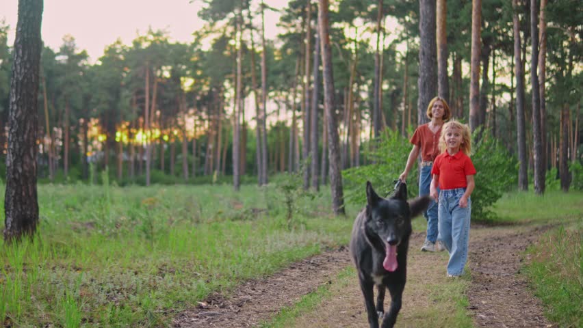 Mother and daughter are walking in the forest and their dog is running in front of them. Cheerful Caucasian family and casual clothes is spending weekend outdoors with a pet. Taking care of animals