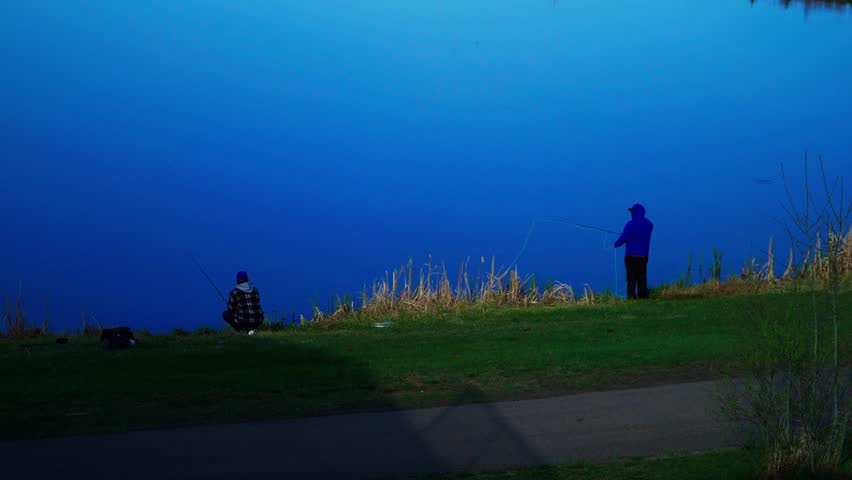 People fish peacefully in a scenic lake under bright morning light. The water reflects vivid blue tones and natural surroundings. Faces are not visible or captured from afar.
