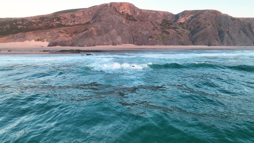 High angle aerial of Praia da Cordoama with rolling waves along cliff-lined beach in Portugal