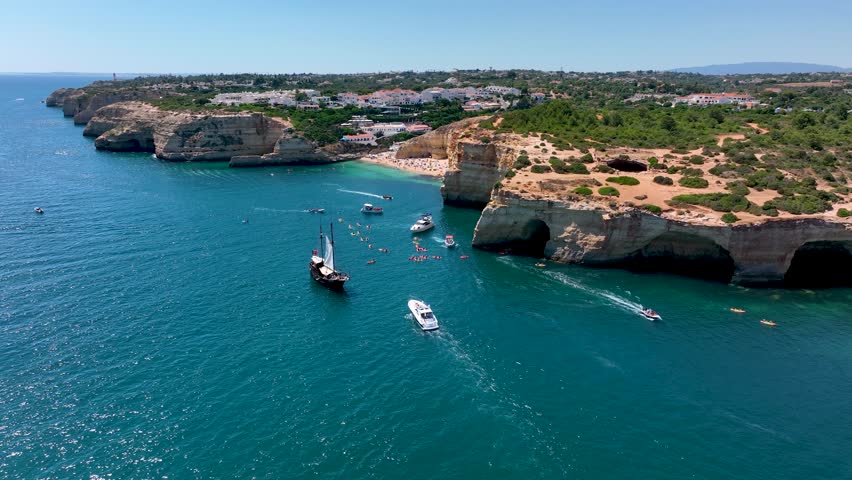 Aerial view of Benagil Cave from the sea with a large group of kayakers, boats, sailboat, and the cave’s roof hole visible along the Algarve coast in Portugal
