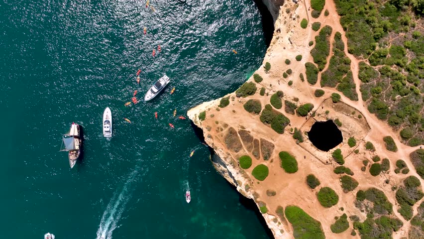 Top-down aerial view of kayakers and boats gathering near the cliffside entrance of Benagil Cave, with the cave’s roof hole visible above in Algarve, Portugal