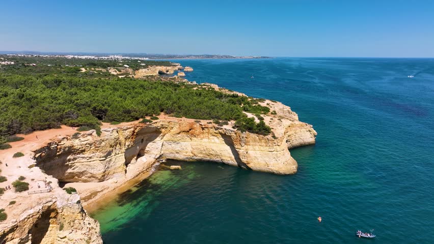 Panoramic aerial view of Algarve’s dramatic coast with sea cliffs, beach coves, boats, forested land, and inland villages under a clear blue sky in southern Portugal