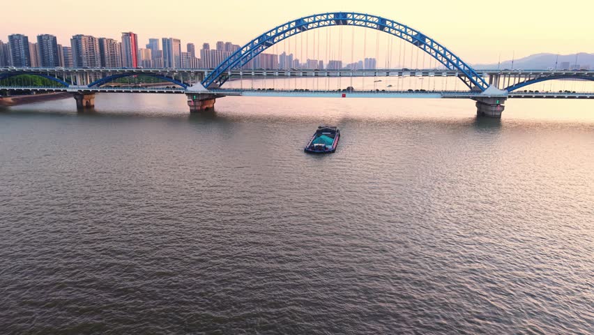 Barge navigating under a modern blue bridge at sunset with city skyline in the background. Hangzhou, China.