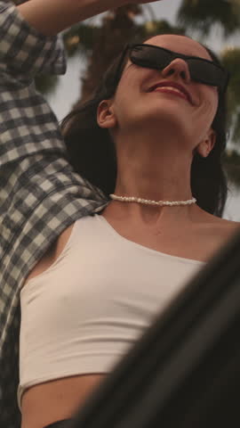 Vertical: Young woman performs dance movements leaning out of car window traveling along road, palm trees. Relaxed girl in sunglasses with pleasant smile revels in tranquility of moment, summer day