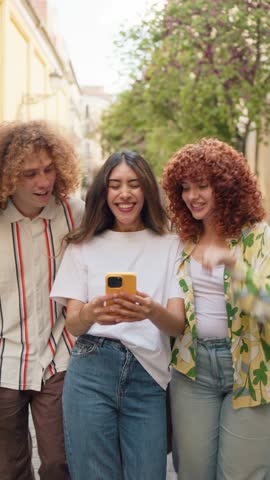 Happy friends looking at smartphone and laughing while walking down the street