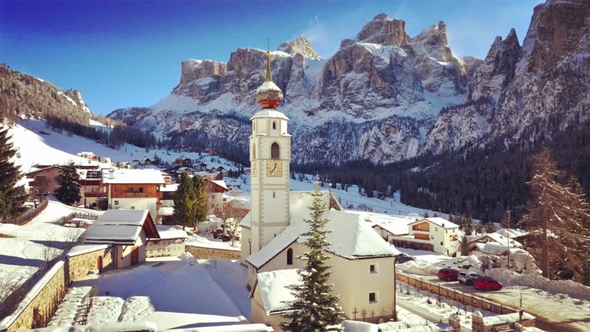 A snow-covered alpine village with a tall church steeple sits beneath majestic mountains under a clear blue sky in a serene winter scene