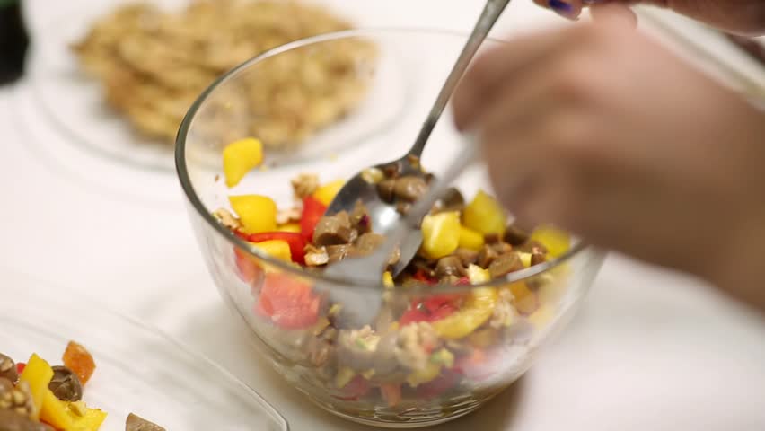 Close-up of hands tossing a vibrant, healthy salad. Chopped red and yellow bell peppers, mushrooms, and walnuts are mixed in a clear glass bowl. Homemade food preparation.