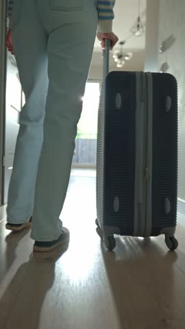 Low-angle view of traveler walking towards door with rolling luggage. Person walks across hallway with orange suitcase. Concept of departure and leaving home for vacation