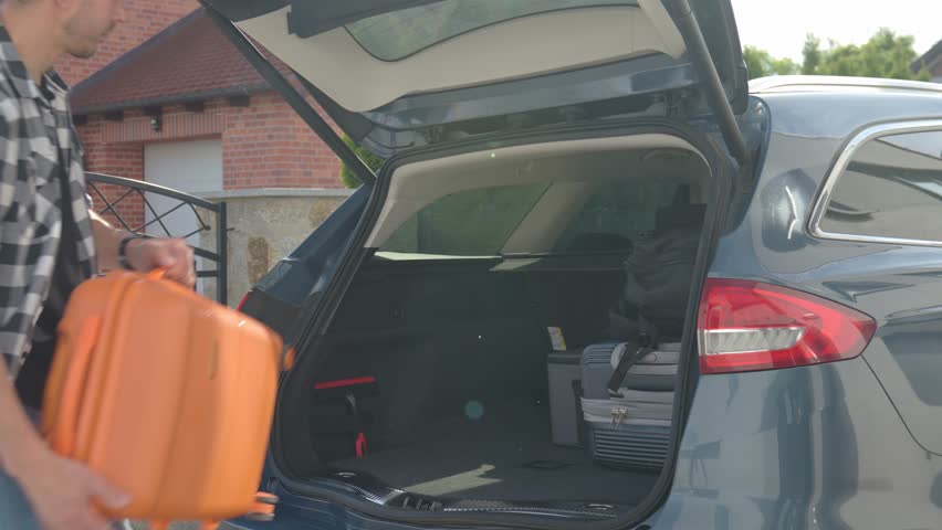 Man loading orange suitcase into car trunk before journey. Packing luggage for travel