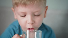 Close up of little boy with blond hair drinking water from clear glass, sitting on bed in cozy home setting, wearing blue shirt, focused on hydrating, refreshing moment of childhood - Powered by Shutterstock - Get 15% off with code: PIKWIZARD15