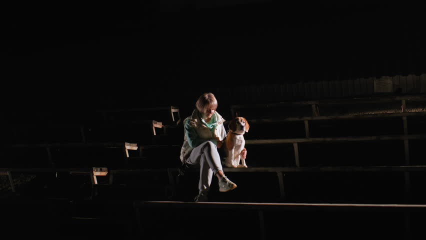 Sports woman seated on wooden stairs at night gently holding german shepherd puppy by her side, illuminated by spotlight in dark empty outdoor stadium creating peaceful and intimate atmosphere