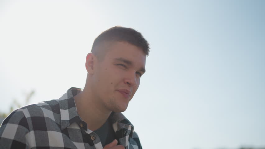 handsome man adjusts checkered shirt while smiling warmly under bright sky with sunlight creating radiant halo effect around head against blurred urban background in casual outdoor setting