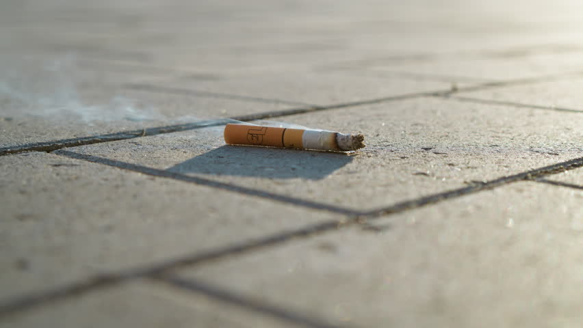 cigarette lying on concrete pavement emitting faint smoke under sunlight, symbolizing addiction, pollution, or abandoned habit with close up perspective and warm ambient lighting