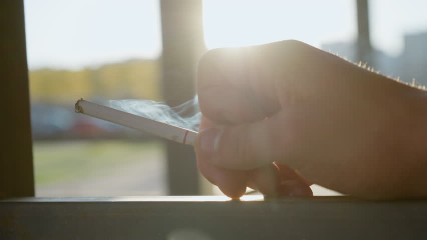 close up of person gently holding cigarette between fingers while smoke swirls upward illuminated by warm sunlight outdoors near railing in soft blurry background during daytime