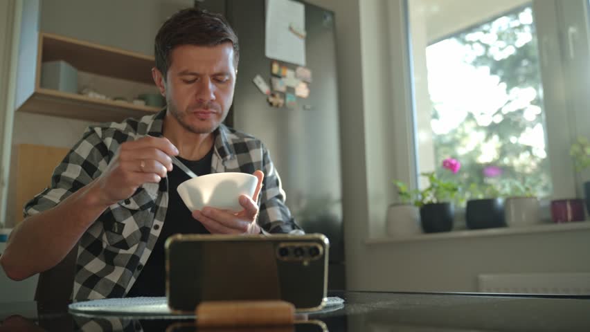 Man in plaid shirt eating oatmeal porridge from white bowl while using smartphone. Male looking at phone during breakfast. Concept of social media addiction, distraction, and digital dependency