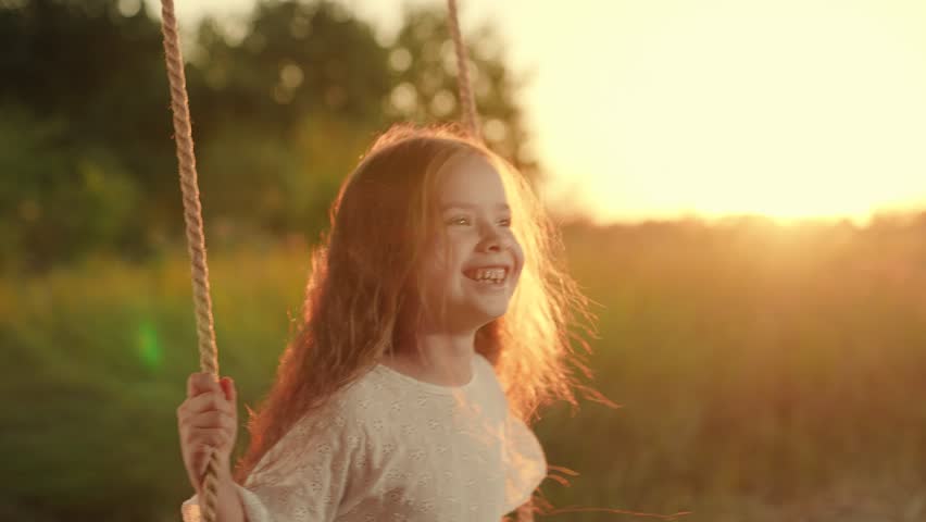 Cute girl child smiles plays on swing, dreams. Child swings on tree on swing. Child's happiness. Child has fun in park in summer. Girl swings on swing in park under tree at sunset. Family walk autumn