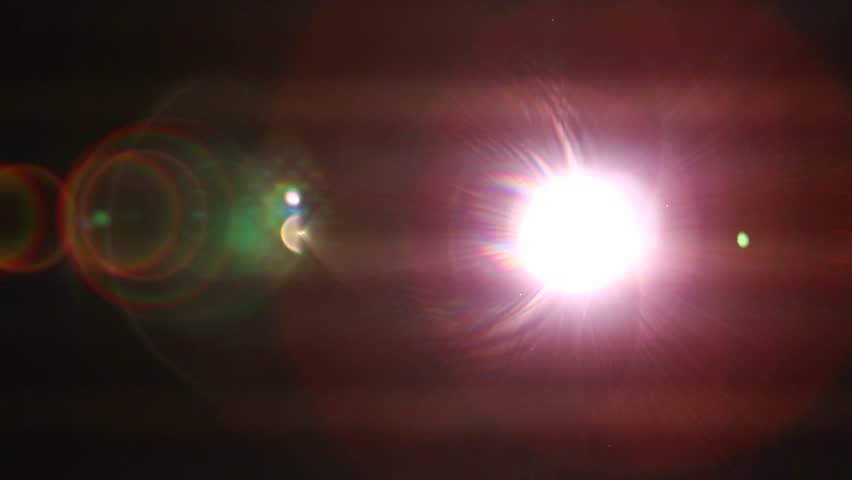 Close-up of a movie projector lens emitting bright, flickering light. Intense red and blue lens flares illuminate dust particles as abstract images project through the lens.