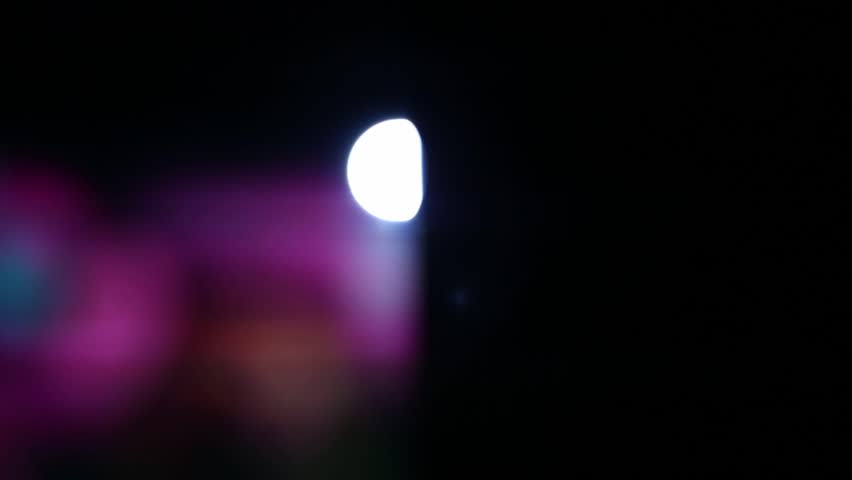 Close-up of a movie projector lens emitting bright, flickering light. Intense red and blue lens flares illuminate dust particles as abstract images project through the lens.