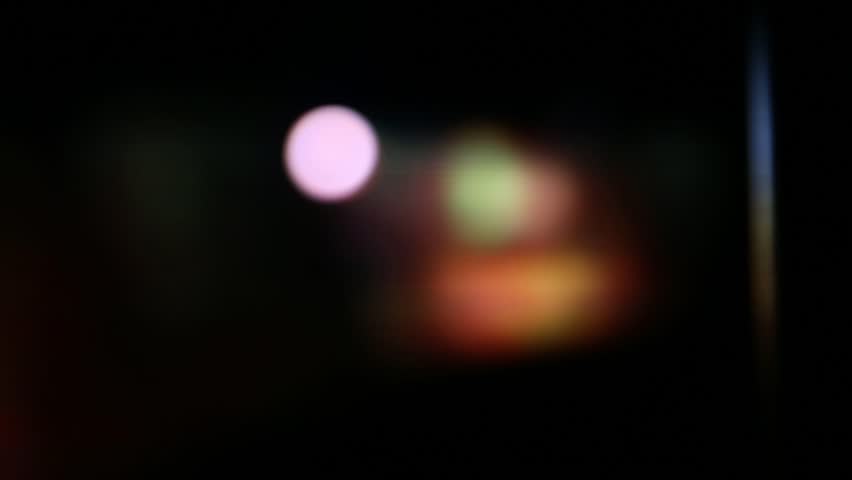 Close-up of a movie projector lens emitting bright, flickering light. Intense red and blue lens flares illuminate dust particles as abstract images project through the lens.