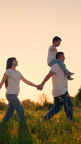 Family walking in park on green grass holding hands, sunset. Happy parents child son on dad's shoulders walking together, sun, nature. Family picnic with child in nature. Dad mom son walking traveling
