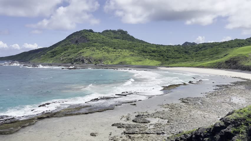 Turquoise blue sea beach, waves movement, green vegetation mound in the background, rocky shore, blue sky, clouds passing, birds. View of Caieira Beach (Praia da Caieira), Fernando de Noronha, Brazil.