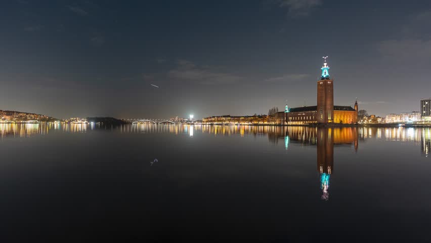 Nighttime timelapse of Stockholm City Hall reflecting on the calm waters of Riddarfjärden with glowing city lights under a clear sky.