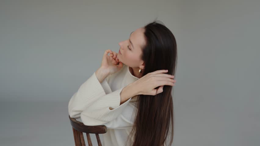 A video of a woman calmly sitting in a chair with her hands placed on her neck. Beautiful young brunette woman with white sweater and pants posing at studio.