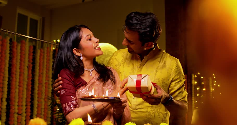 Husband gifting wife during Diwali celebration, Indian couple in traditional attire gifting and placing oil lamp on wooden railing at home, enjoying festive evening with glowing diya and decorations