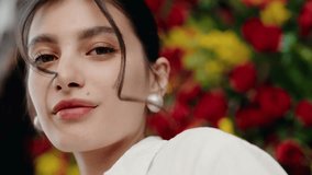 Elegant portrait of a woman with dark hair, wearing pearl earrings, posing in front of vibrant red and yellow flowers. Confident Woman Posing with Vibrant Floral Background - Powered by Shutterstock - Get 15% off with code: PIKWIZARD15