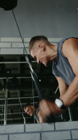 A focused man performs an intense weight training exercise on a cable machine in a modern gym. His determined expression and muscular physique reflect dedication to fitness and training. Vertical