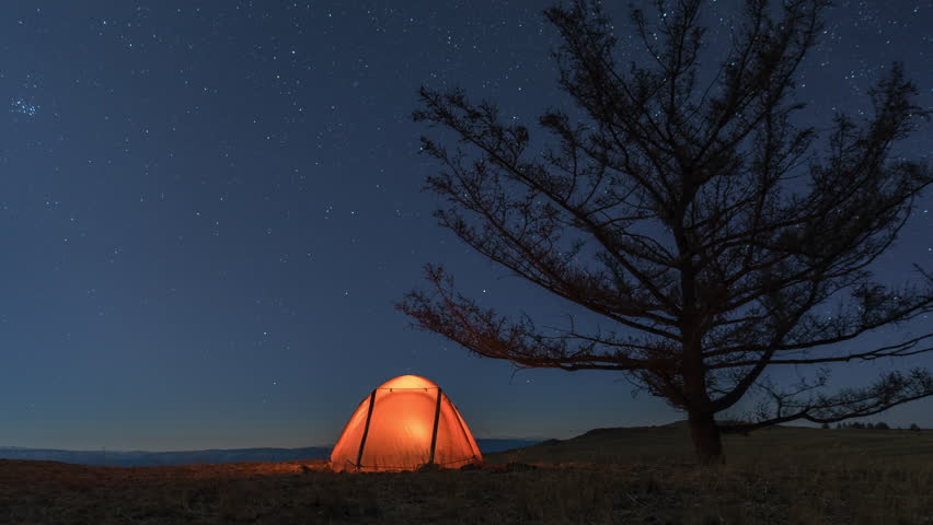 Timelapse of camp with orange illuminated tent near tree at night and moonset