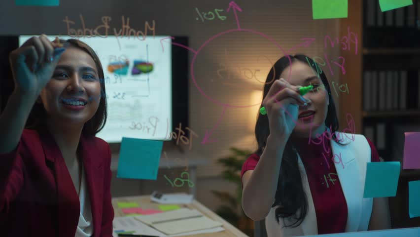 Two women are drawing on a whiteboard with markers. One of the women is smiling. The whiteboard has a diagram on it