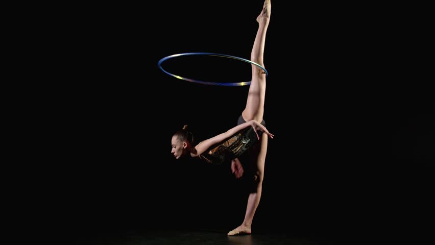 Professional flexible pretty gymnast in elegant leotard practicing rhythmic gymnastic elements, stretching in standing side split with hoop rotating around leg against black background.