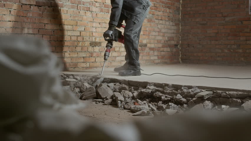 Low angle view of male construction worker wearing hardhat and respirator destroying floors with demolition hammer inside unfinished room