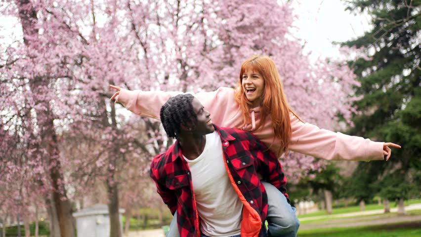 Playful multi ethnic couple enjoying piggyback ride, laughing amid pink cherry blossoms in vibrant spring park setting