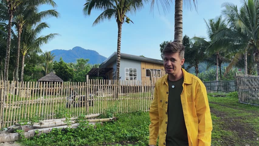Young man travel blogger stands near gate of charming bungalow in Philippines. Male tourist shares impressions of place with genuine excitement