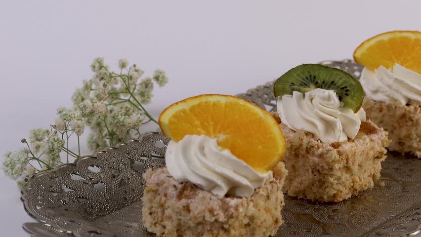 Studio-shot 4K video of two mini fruit-topped cakes with whipped cream, garnished with orange and kiwi slices, placed on an ornate tray. Designed for bakery presentations and dessert promos.
