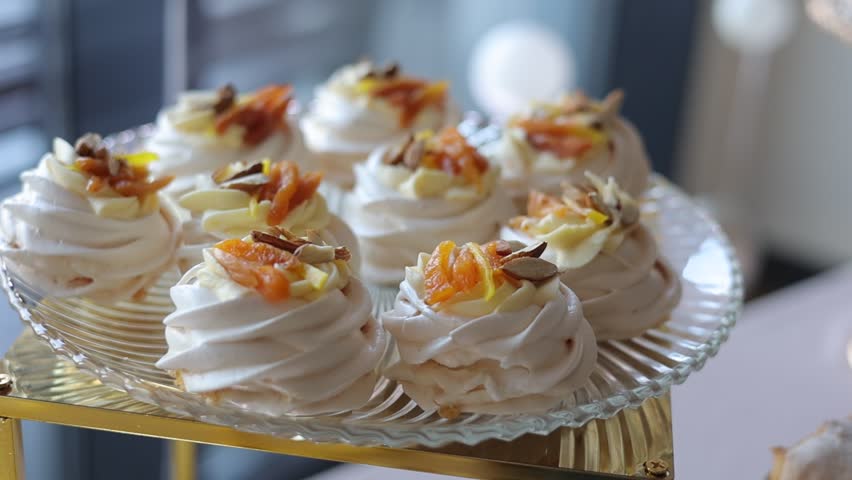 Mini meringue pavlova desserts filled with whipped cream and powdered sugar, decorated with berries, fruits and nuts. Festive sweets served on glass plate and gold stand. Wedding, birthday celebration