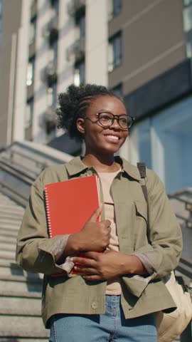 Positive African American student with glasses holding red notebook and smiling while standing outside. Wearing backpack and casual outfit. Heading to university or preparing for morning lecture.