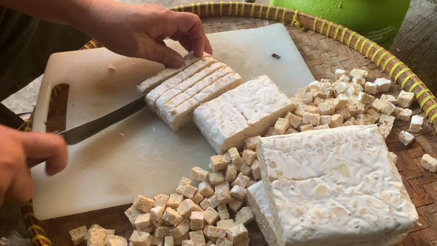 Close-up of hands cutting tempeh into small cubes on a cutting board, traditional Indonesian food preparation scene