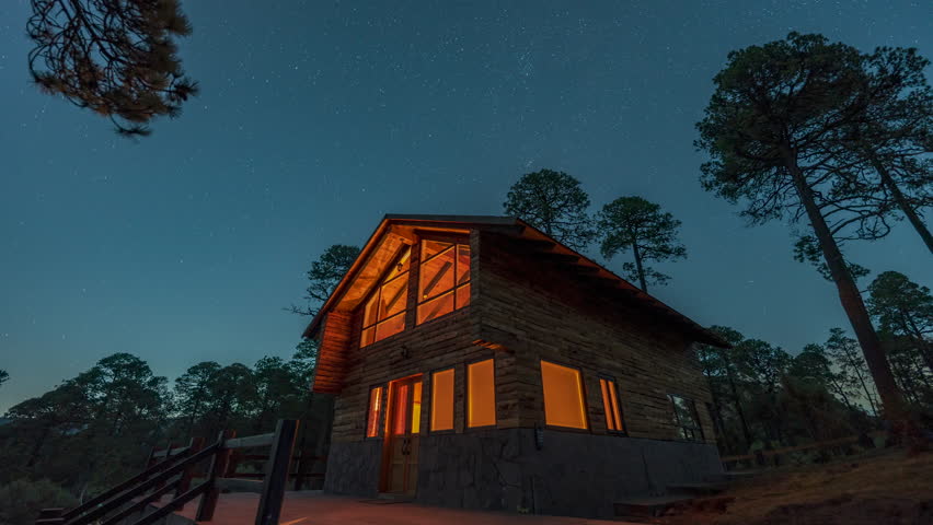 Long exposure of star trails swirling over a warmly lit wooden cabin surrounded by pine trees in a tranquil forest. Timelapse video